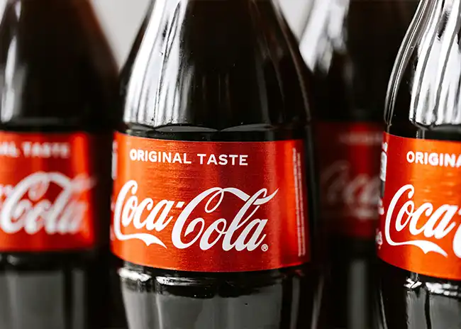 Close-up of several Coca-Cola glass bottles with red labels displaying the iconic 'Coca-Cola' logo and 'Original Taste' text, emphasising the brand's classic beverage.