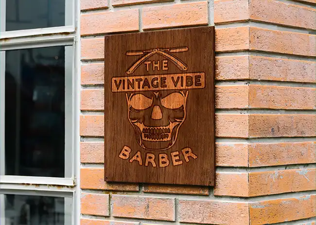 Wooden sign for 'The Vintage Vibe Barber' featuring a carved skull with sunglasses and crossed straight razors, mounted on a brick wall outside a barber shop.