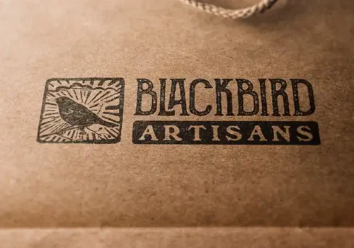 Blackbird Artisans logo featuring a hand-drawn blackbird within a rectangular frame with sunburst rays, accompanied by bold, rustic text 'BLACKBIRD' and 'ARTISANS' on a kraft paper background.