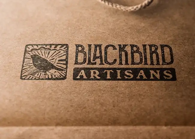 Blackbird Artisans logo featuring a hand-drawn blackbird within a rectangular frame with sunburst rays, accompanied by bold, rustic text 'BLACKBIRD' and 'ARTISANS' on a kraft paper background.