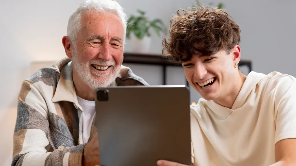 Smiling older man and young man sitting together, enjoying a video or watching content on a tablet at home.
