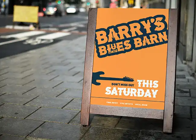 Street sign promoting Barry's Blues Barn, featuring bold, distressed typography on an orange background with a silhouette of a guitar, announcing an event happening 'This Saturday' with details about free entry, live artists, and local brew.