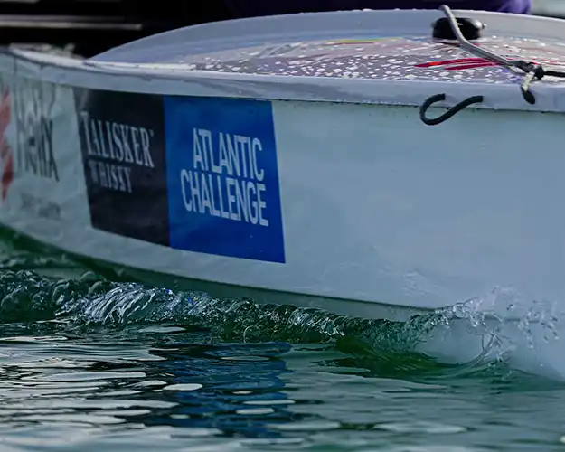Side view of a boat in water, prominently displaying vinyl written sponsors' logos including a whisky brand and an Atlantic challenge event, with waves lapping against the hull