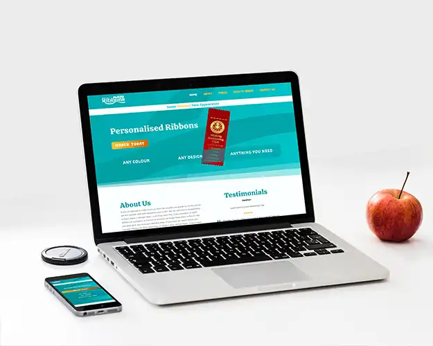 A laptop and smartphone displaying a responsive web design for a personalised ribbons service with a blue and white theme, placed on a minimalist desk alongside an apple and a small device.