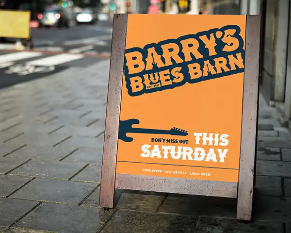 Street advertisement on a sidewalk A-board for a music venue, featuring bold typography and a guitar graphic on an orange background.