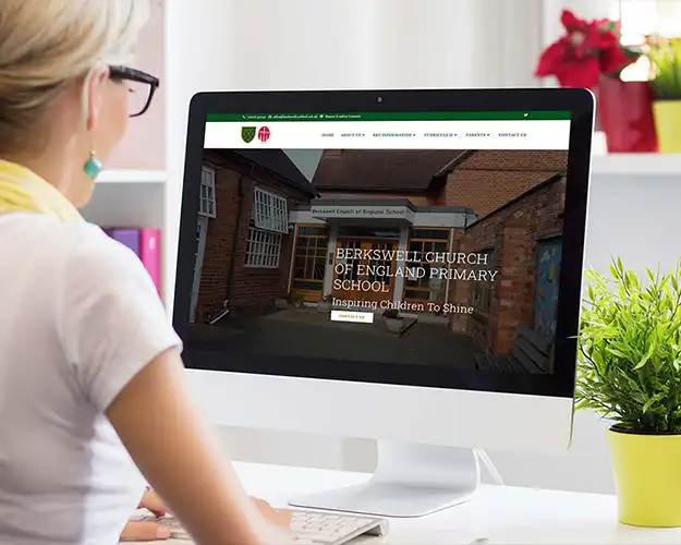 Home office setup featuring a laptop displaying a school's website with the homepage visible, showcasing the school building and its motto. The setting includes a cactus in a white pot and a peaceful, well-lit room.