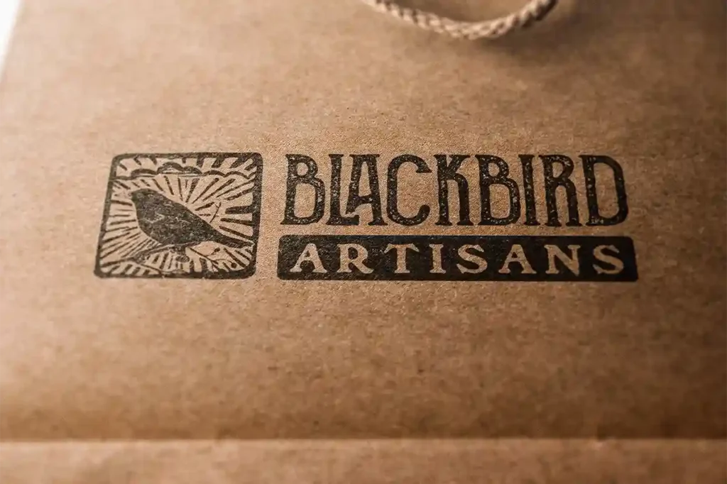 Close-up of a brown paper bag with a vintage stamp logo featuring a blackbird in a decorative square, next to bold text naming an artisan company.