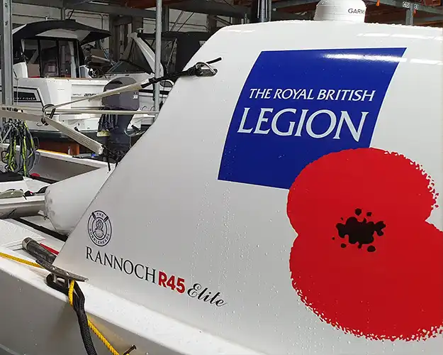Side view of a boat displaying a vinyl printed charity logo alongside a prominent red poppy symbol, docked in a boatyard.