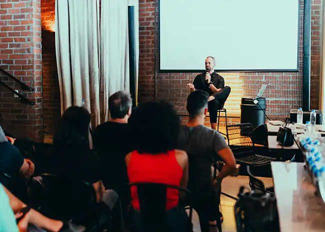 Speaker sitting on a stool and addressing an audience in a brick-walled venue, with attendees seated and listening attentively, creating an intimate and engaging presentation environment.