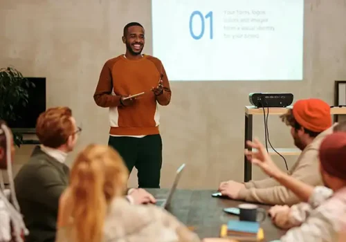 Man giving a presentation to a group of colleagues in a casual office setting, standing in front of a projected slide while holding a tablet, with attendees seated around a table engaged in discussion.