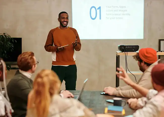 Man giving a presentation to a group of colleagues in a casual office setting, standing in front of a projected slide while holding a tablet, with attendees seated around a table engaged in discussion.