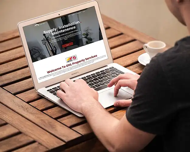 Person using a laptop on a wooden table outdoors, browsing a property services website that highlights services in supply, installation, and maintenance, with a cup of coffee placed to the side.