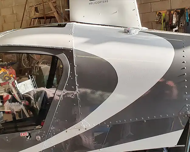 Close-up view of a helicopter's side body showing detailed metallic panels and rivets in a hangar, reflecting a complex, high-precision vinyl wrapping job.
