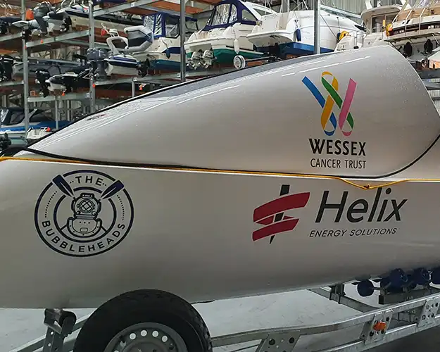 Close-up of a boat's side displaying multiple sponsor logos in vinyl signwriting including a cancer trust and an energy solutions company, parked in a storage facility