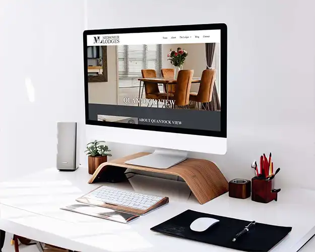 Modern home office setup with a desktop computer displaying a website featuring a luxury lodge. The workspace includes a sleek white desk with stylish accessories like a wood monitor stand, a small plant, and office supplies.