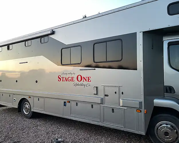 Side view of a large silver horse logo vehicle branded with an upholstery company logo displayed on the body, parked during dusk.