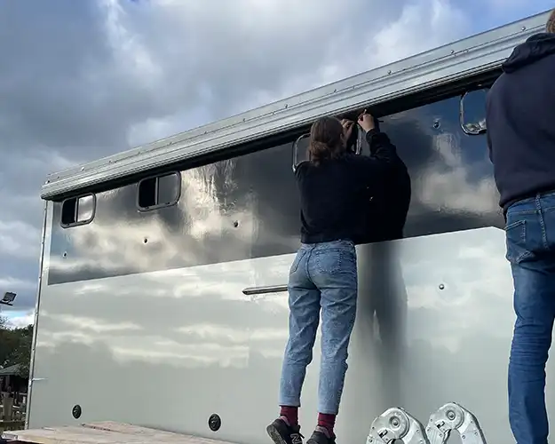 Two people on a scaffolding platform working on vinyl wrapping the exterior of a large silver horse lorry under a cloudy sky.