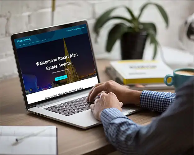 Man in a checkered shirt working on a laptop at a desk, viewing a real estate website. The environment is cozy with a cup of coffee and a green plant on the desk, enhancing the comfortable office setting.