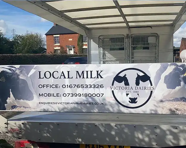 Rear panel of a delivery truck featuring a large promotional banner with images of cows and branding for a local milk service, including contact details.