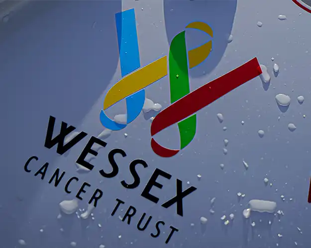 Close-up of a charity organisation's logo with vibrant ribbon-like figures in blue, yellow, red, and green in vinyl graphics on a water-splattered boat, representing support for cancer awareness.