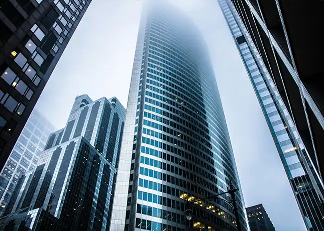 Low-angle view of modern skyscrapers in a city, with the tallest building disappearing into the foggy sky, emphasising urban architecture and atmosphere.