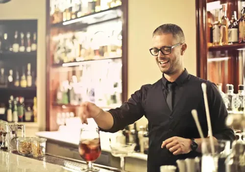 Bartender wearing glasses and a black uniform, smiling while preparing a cocktail at a well-stocked bar, creating a welcoming and professional atmosphere.