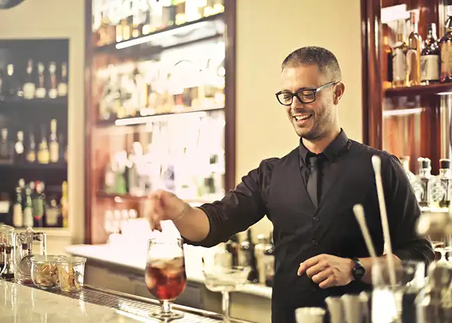 Bartender wearing glasses and a black uniform, smiling while preparing a cocktail at a well-stocked bar, creating a welcoming and professional atmosphere.