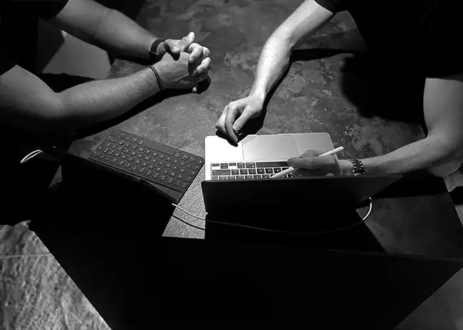 Black and white overhead shot of two people working together at a table, one using a laptop with a stylus and the other with hands clasped, suggesting collaboration or a design discussion.