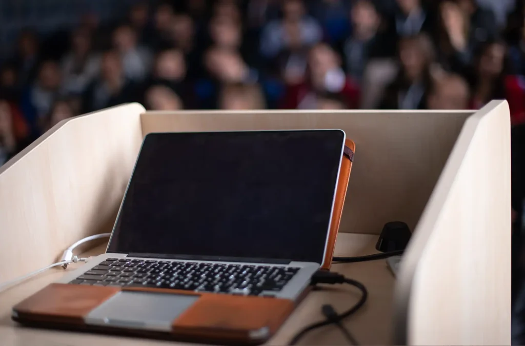 Event graphic design imagery showing a laptop on a podium with a blurred audience in the background, symbolising conference or keynote setups.