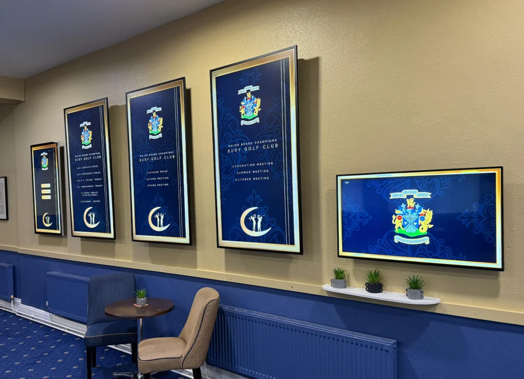 Framed digital display boards mounted on a clubhouse wall, showcasing a series of elegant event and tournament graphics in a refined blue and gold theme.