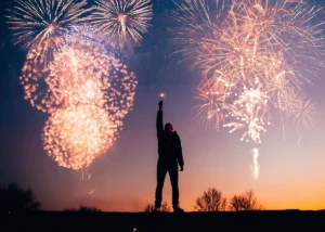 Silhouette of a person holding up a sparkler against a backdrop of colourful New Year’s fireworks at sunset.