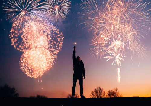 Silhouette of a person holding up a sparkler against a backdrop of colourful New Year’s fireworks at sunset.