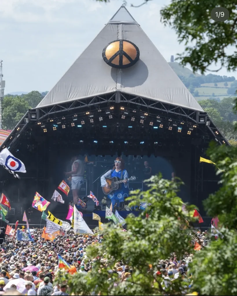 Large outdoor festival stage with a pyramid-style roof and central symbol, live band performing to a packed crowd, with colourful flags waving and greenery framing the scene in bright daylight